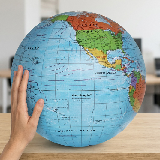 Replogle Inflatable 16-inch Globe (16001) shown from the front at 3/4 angle next to an average-sized hand to provide clear scale reference in a brightly lit home office. The vinyl texture and distinct cartography from the canonical image (image_0.png) are accurately represented.