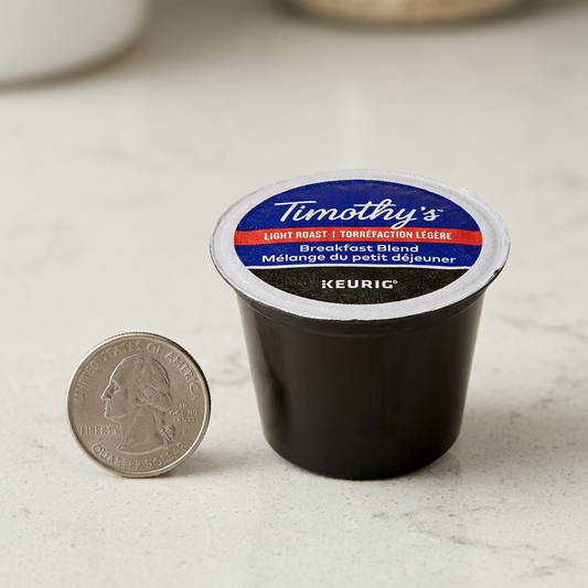 A close-up photograph comparing a standard-sized single-serve coffee K-Cup (Colombian La Vereda) from image_0.png with a standard US quarter, demonstrating its compact size.