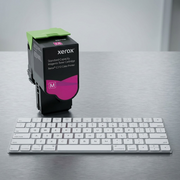 Xerox 006R04358 magenta toner cartridge with green latch standing next to an office keyboard for scale in a workspace.