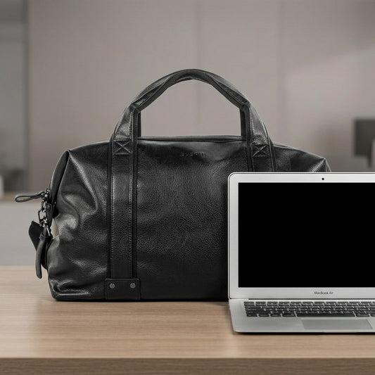 Bugatti Valentino black leather weekender bag next to a 13-inch laptop for size reference on a wooden desk.