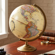 A 12-inch antique ocean desk globe sit on a rich wooden desk with antique-finish books and a classic inkwell pen set, with warm library-style lighting and a brick wall background.