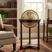 An inviting study features the Replogle Westminster floor standing globe as its central piece. Natural light from a window illuminates the antique-style, French-labeled globe and its classic dark wood stand on a Persian rug. In the background a bookshelf, desk with papers, and armchair contribute to a warm atmosphere. This image perfectly showcases the globe's realistic scale and timeless elegance.