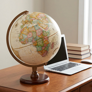 Replogle Hastings globe (30cm/12") placed on a modern wooden desk next to a standard 13" silver laptop, illustrating true size, with natural side lighting.