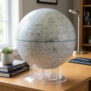 A photograph of the detailed Moon globe on its clear acrylic stand, placed within a contemporary home office environment. The globe sits on a clean, light-colored wooden desk next to a stack of astronomy books and a small succulent plant, with neutral sunlight streaming in from a window. There are no people visible. The globe itself is rendering-accurate to the image_0.png reference.