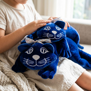 A close-up photograph shows the two-piece fdmt manimo Weighted Cat plush lying across a child's lap in natural light, with soft, checkered bows and detailed embroidery visible.