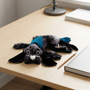 A close-up photo of the manimo weighted dog plush on a wooden shelf next to an average-sized apple.