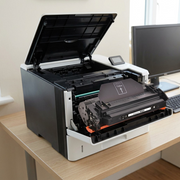 A black laser toner cartridge installed inside an open HP printer in a realistic, lit office setting.
