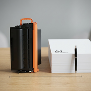 A real-scale comparison view of the Elevate Imaging laser toner cartridge next to an A4 stack of paper and standard pen on a desk.