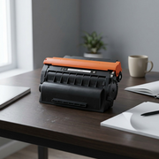 A photograph of an unboxed black toner cartridge with an orange cap on an office desk, illustrating realistic scale.