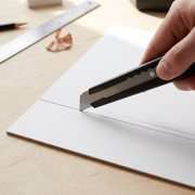 Close-up photo of the OLFA 180 metal knife cutting a straight line on clean white cardboard, on a wooden workbench. (96 characters)