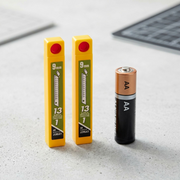 A photorealistic comparison image placing two OLFA 9mm AB-5B yellow blade containers next to a universally recognized standard-sized AA battery to convey the small, intricate scale of the 9mm snap-off blades and their yellow plastic holder.