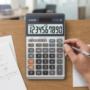 Casio JF-100MS desktop calculator on a wooden desk next to a hand with a pen for scale reference.