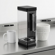 A Keurig K-2500 3.25L clear coffee brewer reservoir with a black lid is shown upright on a stainless steel counter next to a stack of classic paper coffee filters. A standard mug and pen are nearby, clearly demonstrating its considerable size and capacity for a busy environment. Clean product lighting on a white background.