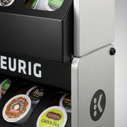Extreme macro view of the STORAGE SHELF-KEURIG's textures: brushed metal side panel, textured black front, clear pod lids, and the iconic Keurig logo, rendered with precision depth of field and premium lighting.