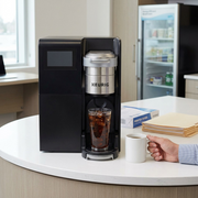 A clean product shot of the coffee brewer beside a hand with a mug for scale in an office breakroom.