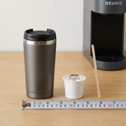A full shot of an accurately scaled Van Houtte Colombian Dark K-Cup coffee pod next to a common wooden coffee stirrer and a medium-sized mug.