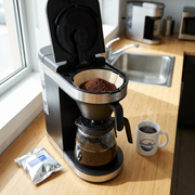 A close-up photograph in a kitchen shows coffee grounds being scooped from an opened silver packet into a coffee filter. The scene features natural morning light, a carafe of freshly dark brew, and a mug in the background.
