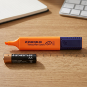 A STAEDTLER orange highlighter is shown next to an ink pen and a hand for scale reference on a desk.