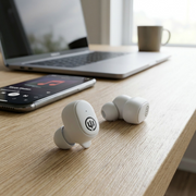 A high-angle shot in a clean home office, featuring the Wicked Audio Embr white earbuds on a desk. They rest on a textured wooden surface next to a smartphone displaying 'Embr Connected' and a closed laptop. Soft daylight enters from a window. Realistic scale, neutral composition. There are no logos or text added to the image.