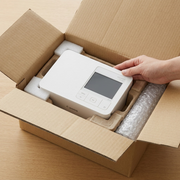 A white Canon SELPHY CP1500 printer is depicted safely embedded within molded protective cardboard packaging inside a neutral corrugated shipping box. The unboxing scene conveys secure transport and careful handling.