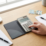 A person uses a silver Canon LS-63TG 8-digit handheld calculator on a light wood desk with a notebook.