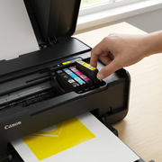 A Canon CLI-281XL Yellow cartridge, without packaging, is placed next to a modern Canon PIXMA printer. A yellow test pattern on white paper is nearby, under natural desk lighting.