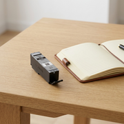 Scale comparison of a single Canon PGI-280 Black Ink Cartridge beside an open journal on a wooden desk.