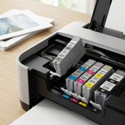 A lifestyle photo shows the specific Canon CLI-221 grey ink tank (from image_0.png) installed within a Canon printer's printhead cradle. The grey tank, clearly marked 'CLI-221 G', is in position alongside other color tanks (C, M, Y, BK). The background is a clean home office desk with soft, natural daylight, including a neat stack of quality photo papers. Focus is on the grey tank in the printer. No packaging. Realistic scale.