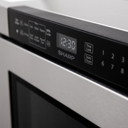 An extreme close-up photograph of the electronic control panel on the SHARP SMD2443JSC Built-In Microwave Drawer. The image shows detailed touch controls, a clear digital clock display showing "12:30", and the prominent "SHARP" logo, all illuminated under premium, low-angle lighting on the matte black interface surface. The depth of field is shallow, highlighting the precise details and reduced purchase hesitation.