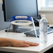 An anti-fog face shield on a desk beside a standard water bottle, with a hand to show scale.