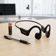 An OS1000BC bone conduction headphone sits naturally on a light wooden desk, positioned adjacent to a standard AA alkaline battery to provide a clear and undeniable sense of its true scale.