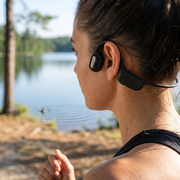 A sporty woman runs on a trail past a ripple on a lake, wearing black bone conduction headphones in natural light.