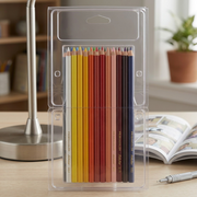 Prismacolor 24-count Scholar Pencils, in their clear blister pack, are shown on a light oak desk next to the base of a silver student desk lamp and the corner of an A4 sketchbook, accurately demonstrating the product's size.