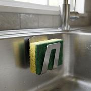 Close-up photorealistic shot of the stainless steel Happy Sinks sponge holder (MPN EB-HS-015) with its black magnetic backing firmly attached to the side of a stainless steel kitchen sink, holding a damp yellow cleaning sponge. Natural kitchen lighting.