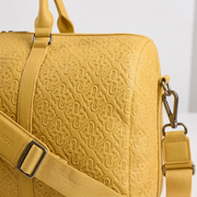 An extreme close-up photograph of the yellow PU leather texture on a Bugatti Rick Ross Edition bag. Detail shot shows the complex embossed repeating logo pattern with precision stitching and a metal swivel hook connector.