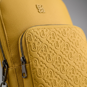 Macro detail of the Bugatti sling bag focusing on the textured yellow leather, embossed logo, and metal hardware.