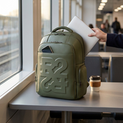 The green leather-like backpack is sitting on a train table, a laptop slightly out, near a window at dusk.