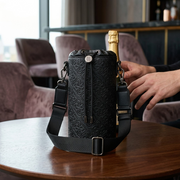 A low-angle, close-up photograph captures the Bugatti Rick Ross Champagne Bag, precisely as depicted in image_0.png with its debossed monogram and leather-textured black PU, resting confidently on a table in a stylish lounge. An out-of-focus hand by an empty crystal champagne flute is visible near it.
