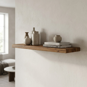 A lifestyle image showing the natural wood floating shelf mounted in a modern living room, holding a vase and a plant.