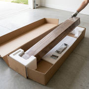 A gloved hand lifts a wrapped walnut mantel beam from custom heavy-duty shipping packaging on a clean floor.