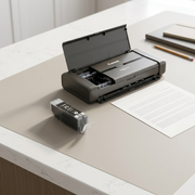 A high-angle photo of a Canon PGI-35 Black ink tank resting on a clean white desk, positioned next to a Canon Pixma iP110 portable printer with its top cover slightly propped open, ready for use, with a blank sheet of paper and a closed notebook in the background. The ink tank label is clearly visible. The lighting is soft and natural. The packaging is not in view.