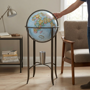 An floor standing 16-inch or 40.5-centimeter diameter globe, with a light-blue ocean, is shown in image 0, positioned in a living room, standing beside a mid-century modern armchair. The arm of the chair is almost exactly the same height as the globe's horizontal metal band.