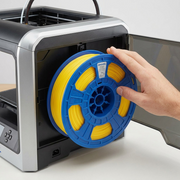 A scale-reference image showing the Dremel yellow filament spool next to an open 3D printer, demonstrating its fit.